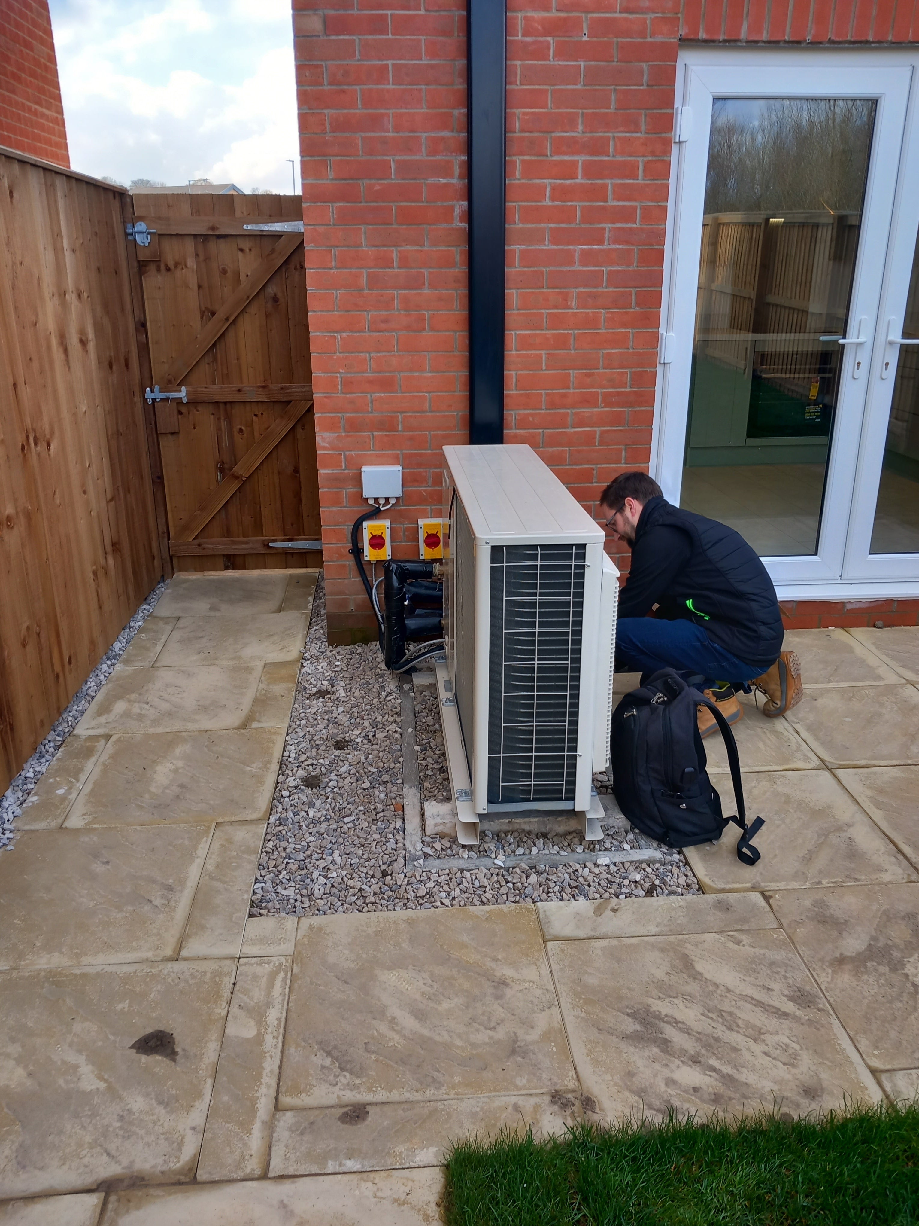 Person working on an air conditioning unit outside a building with a brick wall and wooden deck.